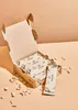 A cardboard box filled with individually wrapped raw cashew bars, with some loose cashews scattered around the box. Produktbild-Vorschau