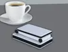 A white coffee cup and saucer are in the upper left corner of the image. To the right of the cup, a stack of two white rectangular objects with black trim and small black wheels is shown. Produktbild-Vorschau