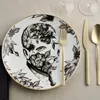 A close-up of a decorative plate with a floral and portrait design, set with gold cutlery and wine glasses in the background. Produktbild-Vorschau