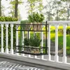 A black metal plant stand with two shelves is hanging on a white railing on a balcony or deck. Several potted plants are displayed on the shelves, and a green, leafy hedge is visible behind the railing. The background shows trees and a grassy area. Produktbild-Vorschau