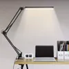 A black adjustable LED desk lamp is positioned on a wooden desk, illuminating a white wall. Produktbild-Vorschau