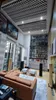 A spacious, modern living room with high ceilings, featuring a large brown leather sofa, two white coffee tables, and several large black speakers. The ceiling has a black and white checkered acoustic treatment, and the walls are adorned with artwork. A ta Náhľad obrázku produktu