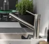 A close-up shot of a modern kitchen faucet with a pull-out spout, set against a light gray and white countertop. A potted herb plant is visible in the background. Anteprima dell’immagine prodotto
