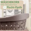 A close-up of a brown woven laundry basket with a wood-grain textured rim, set against a blurred background of a laundry room with a washing machine and shelves. Produktbild-Vorschau
