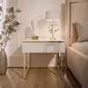 A white high-gloss nightstand with a gold frame and accents, placed next to a bed. A lamp and a bust are on top of the nightstand. A vase with decorative branches is to the left. Náhled obrázku produktu