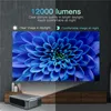 A projector displaying a large blue flower image on a wall, with the projector itself placed on a wooden cabinet in the foreground. Voir les images du produit