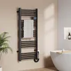 A black electric towel warmer is mounted on a light beige wall in a bathroom, with a white towel draped over it. A plant is visible to the left, and a white bathtub with a wooden tray and a candle is to the right. Anteprima dell’immagine prodotto