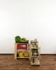 A wooden shelving unit stocked with groceries, including produce, canned goods, and boxed items, against a white wall. Produktbild-Vorschau