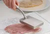 A hand is using a meat tenderizer on a piece of meat covered in plastic wrap. In the background, a piece of breaded meat is on a plate. Podgląd zdjęcia produktu