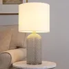 A table lamp with a textured glass base and a white cylindrical shade sits on a marble-top side table next to an upholstered chair. Anteprima dell’immagine prodotto