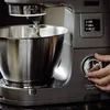 A close-up shot of a person's hand adjusting a dial on a Kenwood Titanium Chef Patissier XL stand mixer. The mixer has a digital display showing timer information. Produktbild-Vorschau