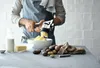 A person is using a potato ricer to mash potatoes into a bowl, with ingredients like milk and butter on the counter. Produktbild-Vorschau