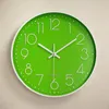 A green wall clock with white numbers and hands is shown against a textured light beige wall. Náhled obrázku produktu