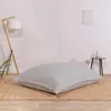 A large, light gray floor cushion with a subtle striped pattern is placed on a woven rug in front of a white wall. A wooden floor lamp with a white shade stands to the left, and a small black side table with a decorative object is to the right. Voir les images du produit