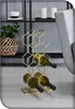 A golden metal wine rack holding three bottles of wine, placed on a floor next to a chair and a shelf with a plant and candles. Produktbild-Vorschau