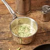 A close-up shot of a stainless steel saucepan filled with a creamy sauce and herbs, placed on a wooden table. Two decorative tin cans are visible in the background. Náhled obrázku produktu