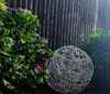 A decorative sphere with lights sits next to a flowering plant in front of a wooden fence. Produktbild-Vorschau