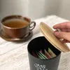 A hand is opening a black container filled with tea or sweetener packets, next to a steaming cup of tea. Náhľad obrázku produktu