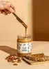 A hand is spreading almond butter from a jar onto a slice of bread, with almonds scattered around. Produktbild-Vorschau