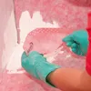 Hands wearing teal gloves are applying a pink waterproofing membrane with white dots to a corner of a wall that has white sealant applied. Náhľad obrázku produktu