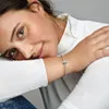 A woman wearing a white shirt and blue jeans, resting her head on her arm, showcasing a bracelet on her wrist. Anteprima dell’immagine prodotto