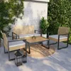 An outdoor patio set with a sofa, two chairs, and a coffee table on a circular rug, surrounded by plants and a textured wall. Produktbild-Vorschau