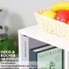 A close-up view of a white shelf unit with books and decorative fruit in a basket. Produktbild-Vorschau