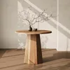 A round wooden dining table with a vase of branches and a small bowl on top, set against a textured wall with light casting shadows. Produktbild-Vorschau