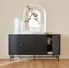 A black sideboard with fluted doors and a metal base, decorated with glass vases and a plant branch, placed in a room with an arched window in the background. Produktbild-Vorschau