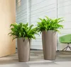 Two tall taupe planters with lush green plants are placed on a tiled floor next to a wooden door and a green chair. Produktbild-Vorschau
