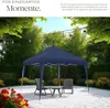 A navy blue gazebo is set up on a paved patio in a garden, with two chairs and a small table placed underneath it. String lights are hung above the gazebo. Náhled obrázku produktu