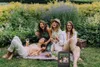 Four women are having a picnic in a garden, laughing and holding wine glasses. A small screen is visible in the foreground, showing a picture of the women. Produktbild-Vorschau