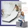 A woman is setting up a foldable mattress in a bright, modern room. Another woman is sitting on a folded ottoman. Produktbild-Vorschau