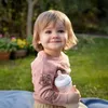 A young child sits on a blanket in a garden, holding a baby bottle. Produktbild-Vorschau