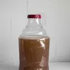 A clear plastic fermentation vessel filled with a brown liquid, with a red cap and a small airlock attached to the neck. Produktbild-Vorschau