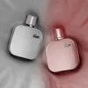 Two perfume bottles, one silver and one pink, are placed on a textured fabric background. Podgląd zdjęcia produktu