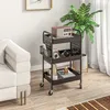 A dark brown rolling cart with three shelves is placed next to a white sofa and a large potted plant. The cart holds various items, including a vintage radio, camera, headphones, and decorative vases. Produktbild-Vorschau