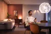 A man is sitting at a dining table in a modern living room, holding a smartphone and looking up at a decorative pendant light. Náhled obrázku produktu