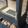 Close-up view of a modern wood-burning stove with a brass handle and a storage compartment filled with firewood. Produktbild-Vorschau