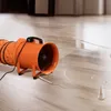 An orange industrial fan with a flexible hose is placed on a wet wooden floor, suggesting it is being used for drying or ventilation in a construction or renovation setting. Náhled obrázku produktu