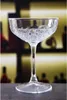 A clear, well-lit studio shot of a single crystal coupe glass, placed on a dark gray surface. The background is blurred, showing shelves with bottles, suggesting a bar or restaurant setting. Produktbild-Vorschau