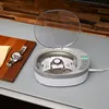 An ultrasonic cleaner with a watch inside sits on a grey surface next to a wooden board with a watch on it. Produktbild-Vorschau