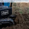 A person's foot is on the platform of a mini excavator, which is digging into the dirt. Produktbild-Vorschau