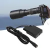 A camera with a long lens is shown in use, with a dummy battery and cable in the foreground. Náhled obrázku produktu