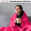 A woman is sitting on a couch wrapped in a pink blanket, holding a mug. Produktbild-Vorschau