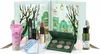 A collection of beauty products, including a makeup palette, lipstick, mascara, face mask, and skincare items, arranged in front of an open advent calendar decorated with a winter village scene. Produktbild-Vorschau