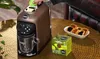 A Lavazza coffee machine is shown on a table with a box of coffee pods and some pastries. Podgląd zdjęcia produktu