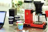 A red Moccacoffee machine with a glass carafe and water reservoir sits on a counter next to a laptop and several coffee cups. Náhled obrázku produktu