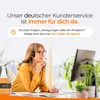 A woman wearing a headset is working at a desk, with a computer monitor and a bottle of cleaning product visible. The background includes plants and a window. Produktbild-Vorschau