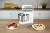 A white stand mixer with a silver bowl is placed on a wooden countertop, with a plate of berries and whipped cream on the left and a plate of cake slices on the right. Anteprima dell’immagine prodotto